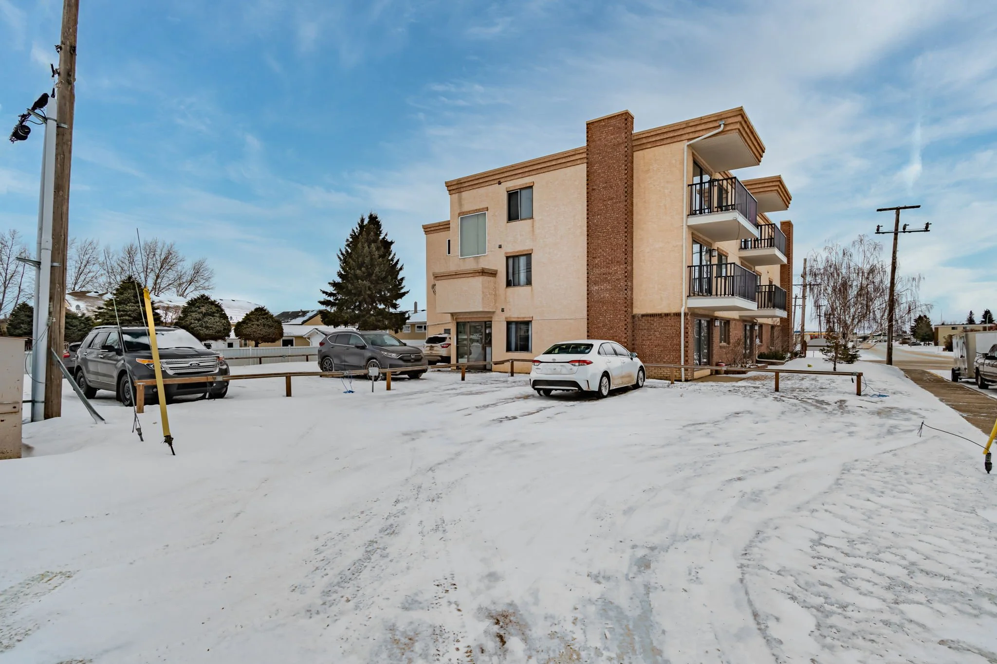 Snow-covered parking lot in front of a three-story building with balconies, featuring several parked cars and scattered trees under a partly cloudy sky.