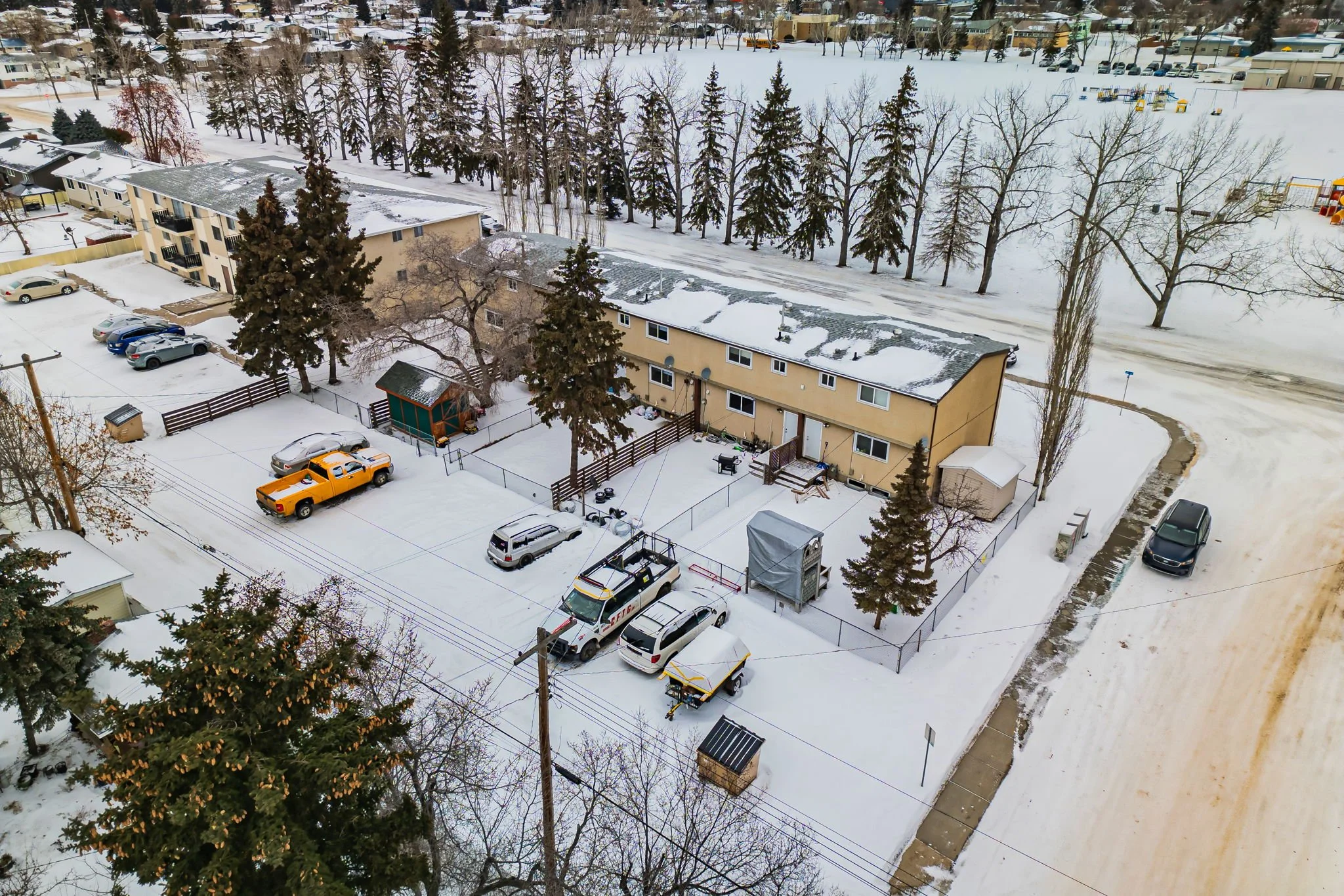 Snow-covered residential area with parked cars, trees, and an apartment building in winter.