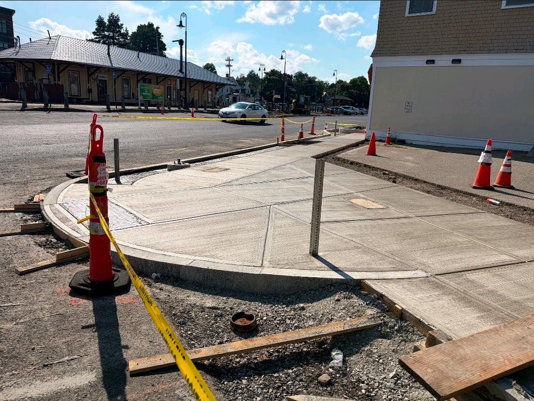 Freshly poured curved concrete sidewalk under construction near downtown Reading's train depot, marked off with cones and caution tape.