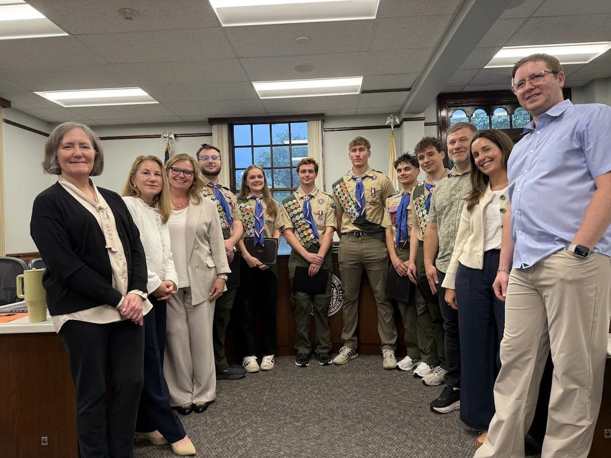 Six Eagle Scouts in uniform, wearing merit badge sashes and holding certificates, stand with members of the Select Board in the meeting room at Reading Town Hall.