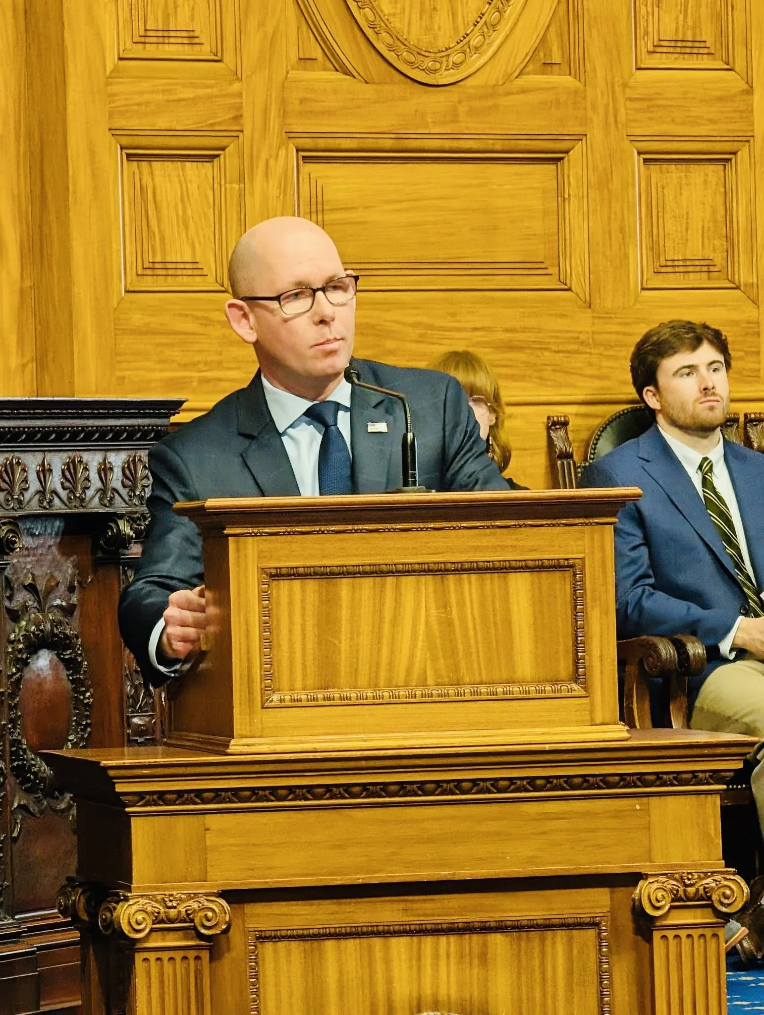 Rich Haggerty, a bald man in a dark suit and blue tie, speaks from a wooden podium in a wood-paneled State House chamber, with two people seated behind him.