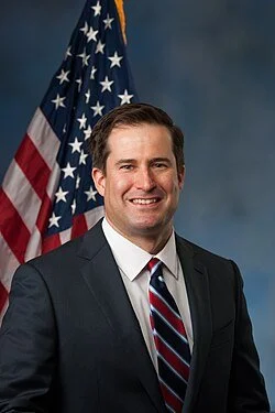 Official portrait of Representative Seth Moulton, a middle-aged white man with dark hair, wearing a dark suit and a red, white, and blue striped tie, standing in front of an American flag.