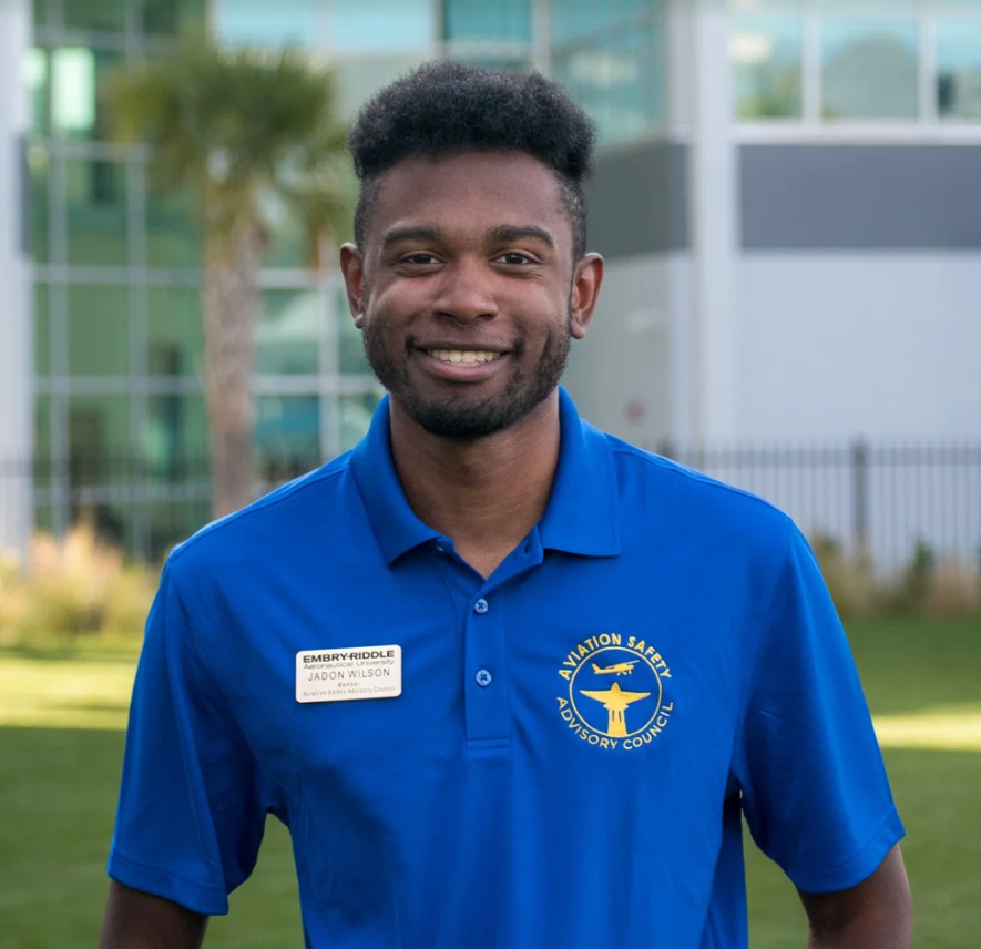 Portrait of Jadon Wilson, a smiling young Black man with a beard, wearing a blue Aviation Safety Advisory Council polo shirt with an Embry-Riddle name tag, outside a glass building.