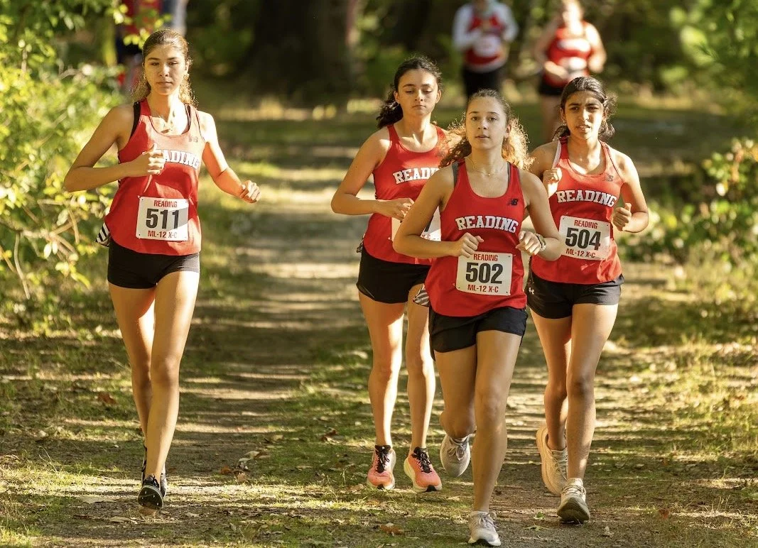 Four Reading girls' cross-country runners in red jerseys run together on a wooded dirt trail during a Middlesex League meet.