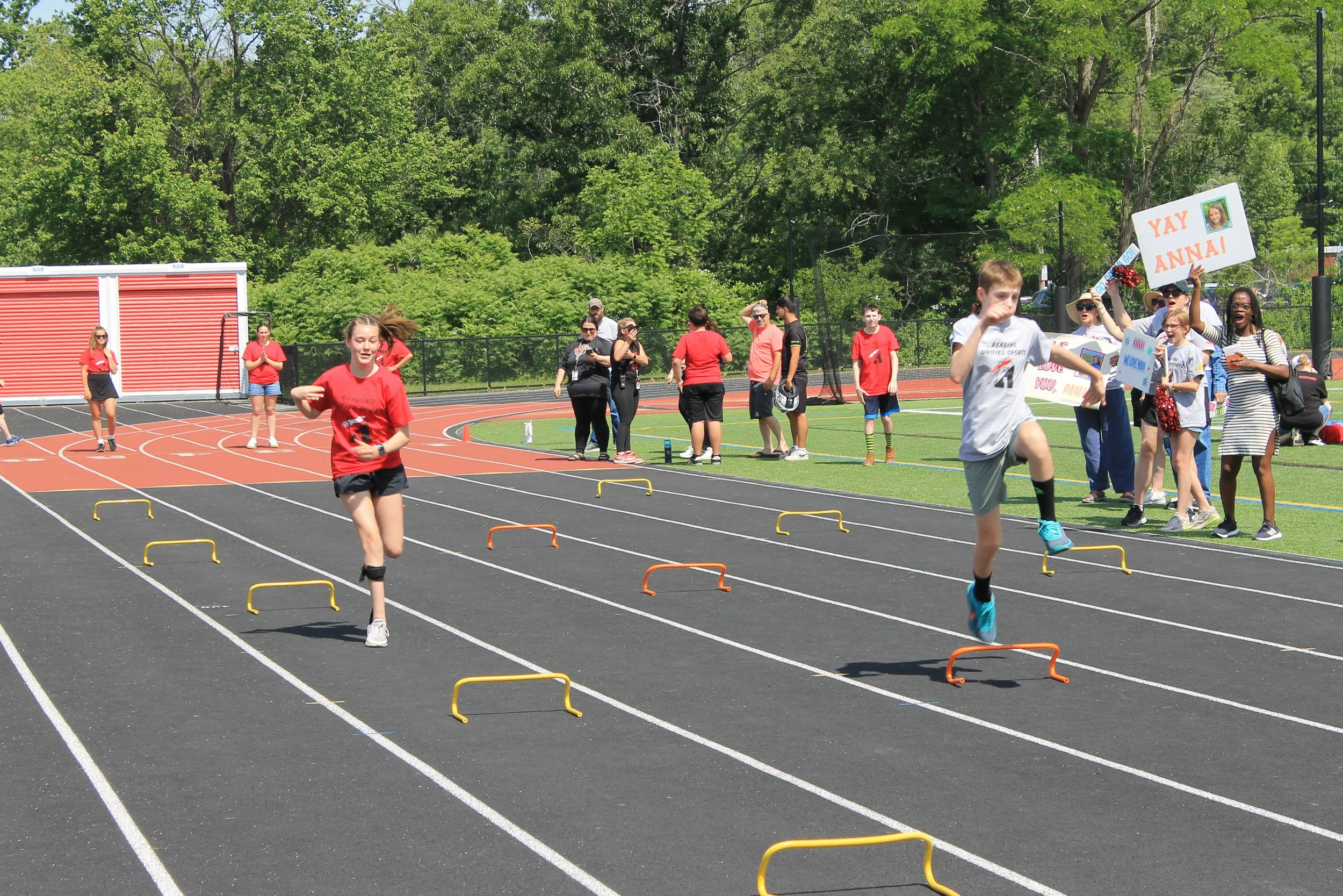 Two athletes clear hurdles side by side as a sign reading "Yay Anna!" cheers them on.