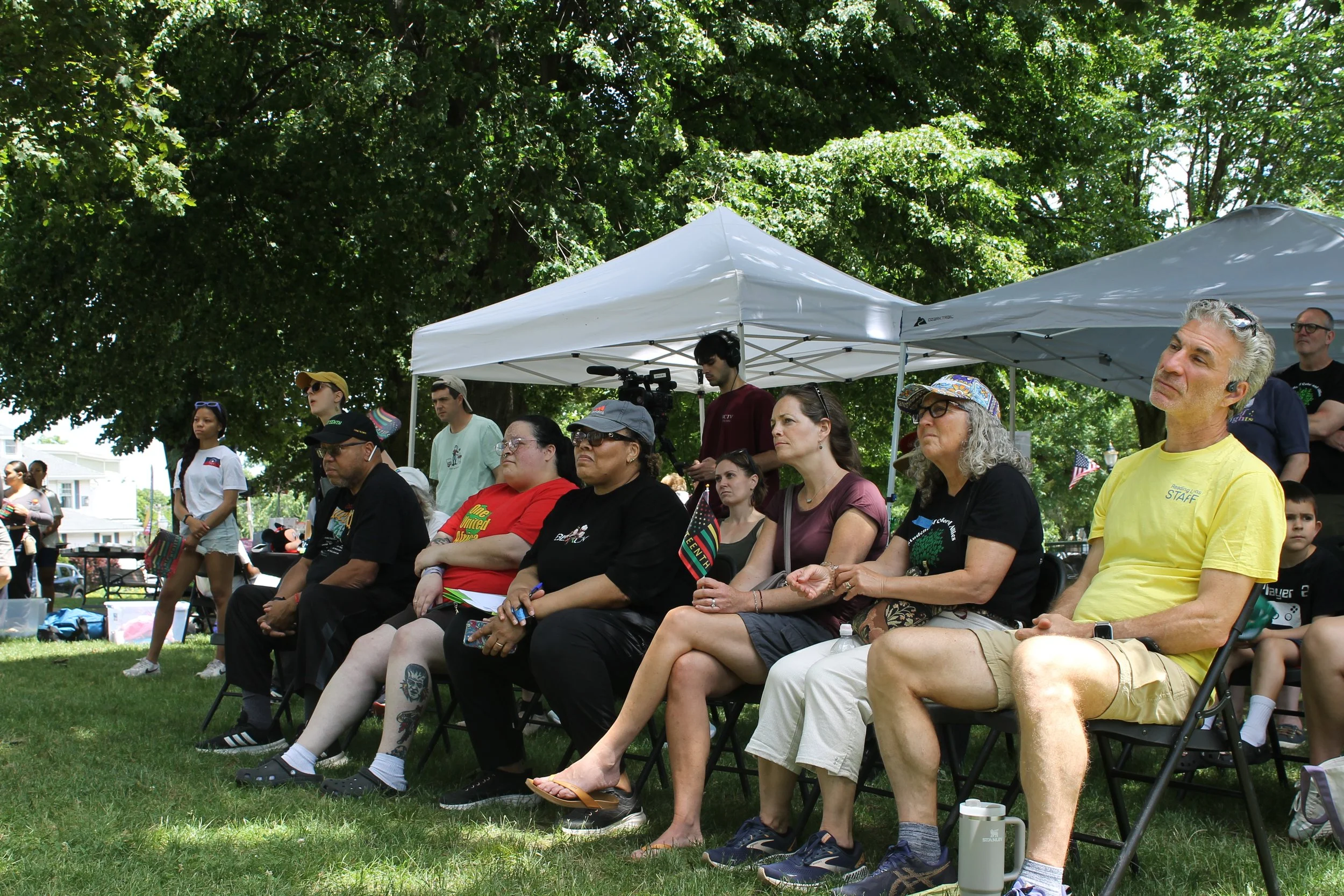 Attendees settle into folding chairs in the shade, enjoying the program.