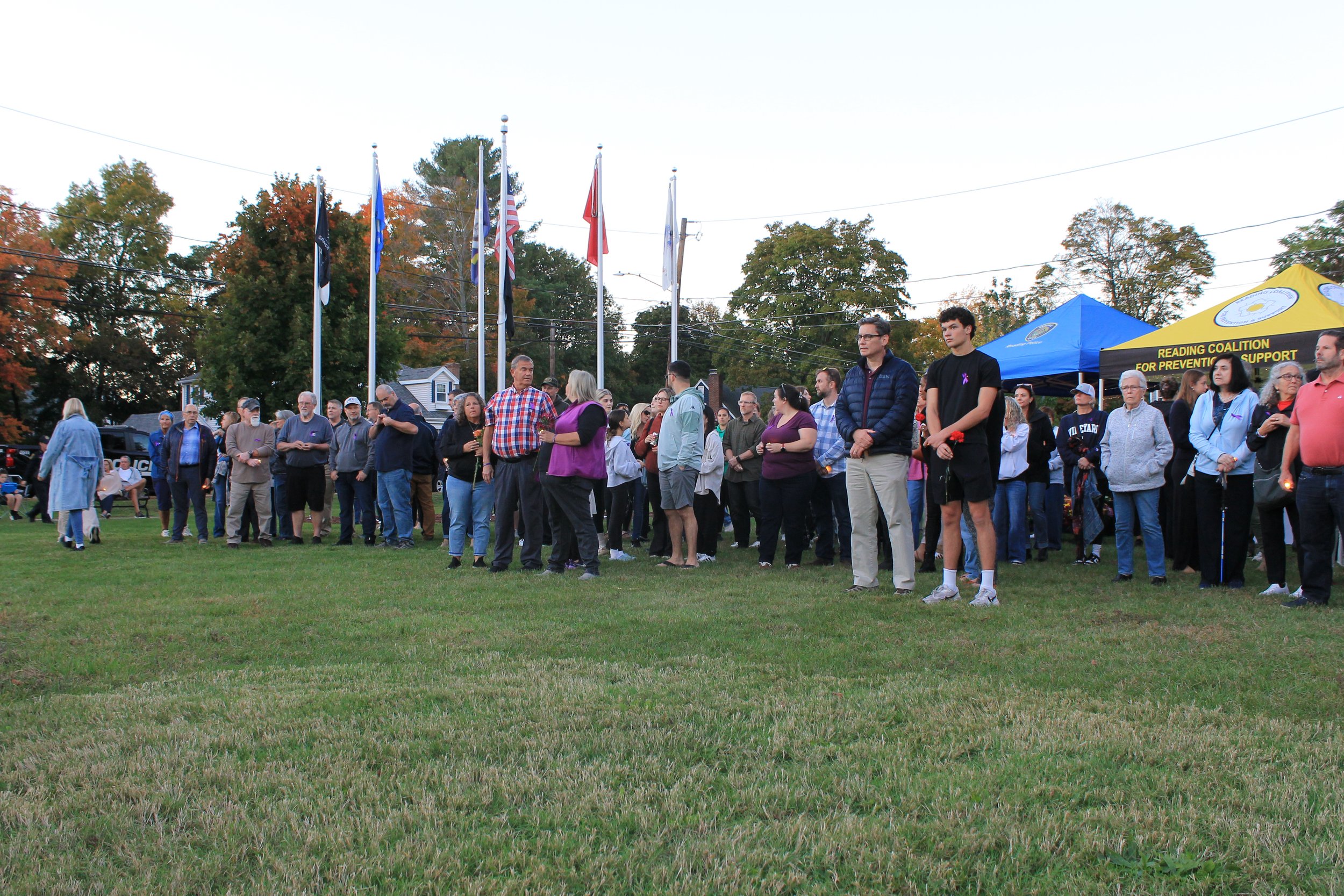 Attendees gather near the flagpoles holding candles and flowers.
