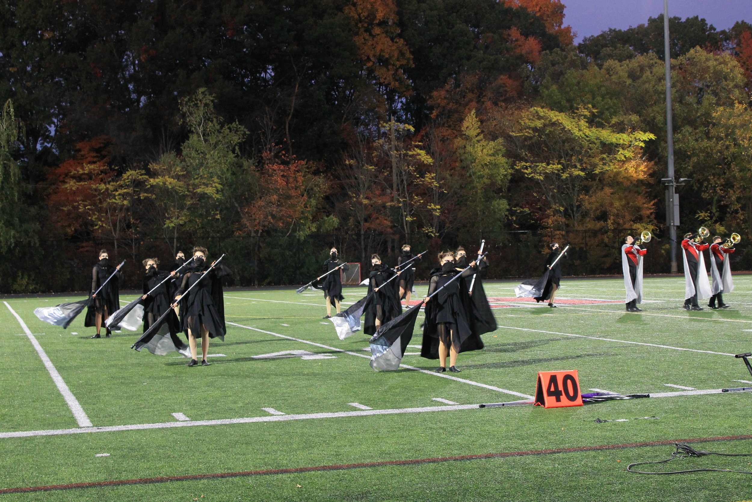 Color guard members in black costumes perform with flags as brass players perform nearby.
