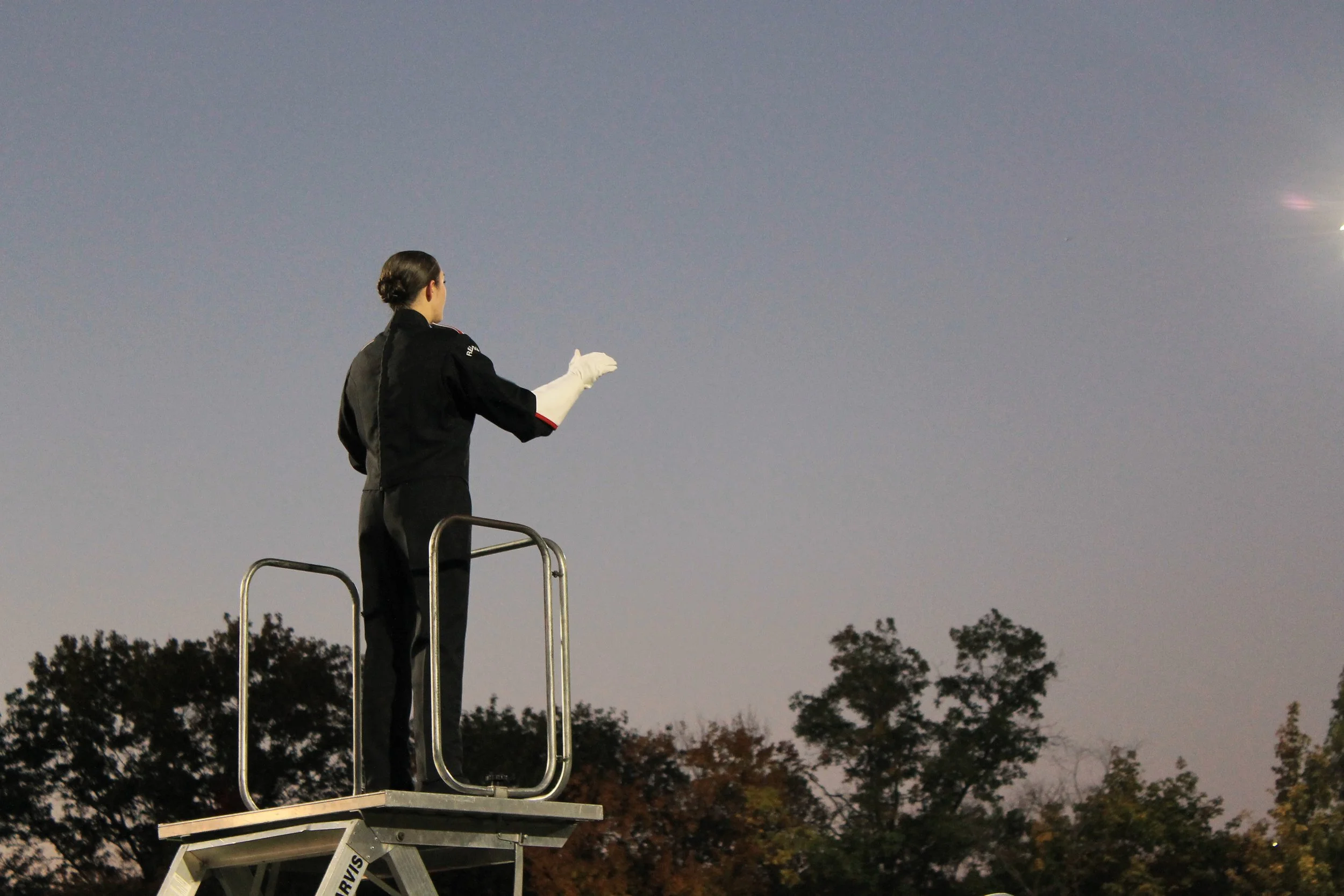 The drum major conducts from an elevated platform as dusk settles over the field.