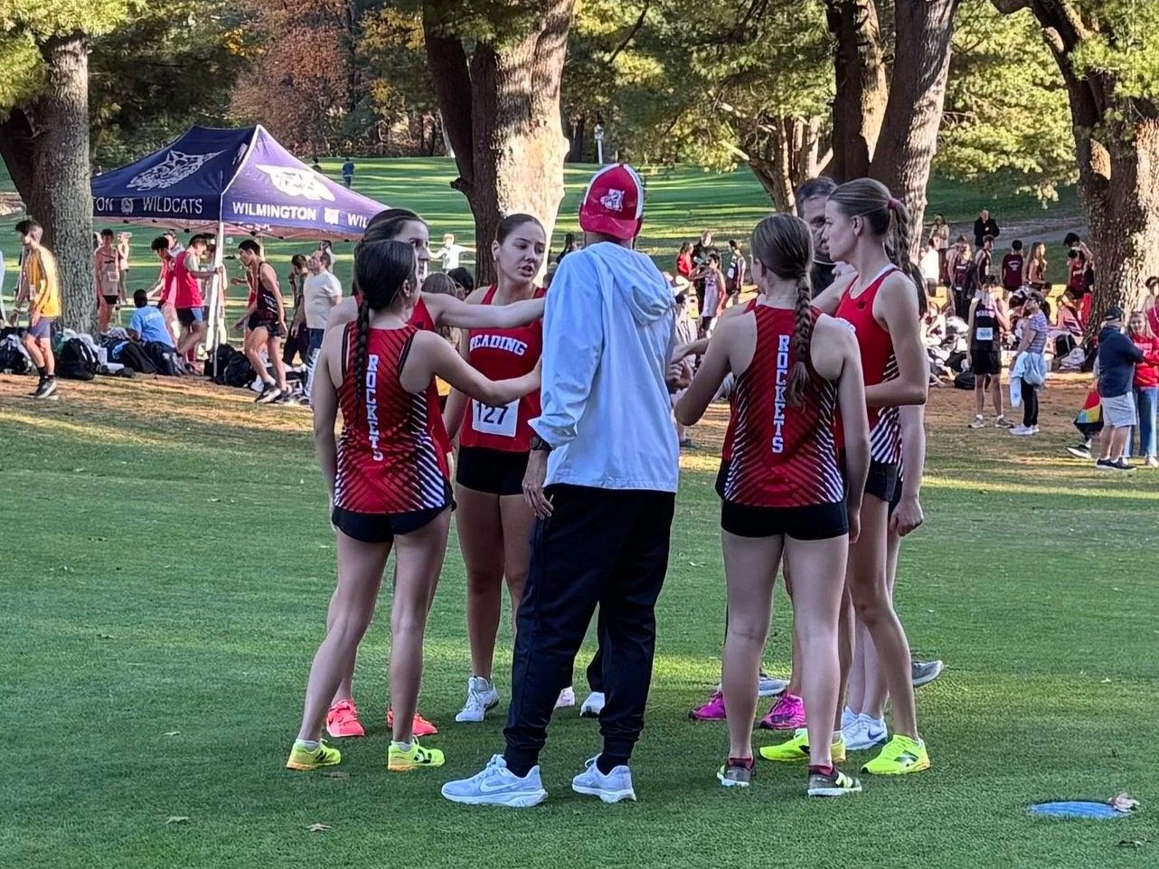 Reading girls cross-country runners in red uniforms huddle around a coach in a white jacket and red cap on a grassy course, with a Wilmington team tent in the background.