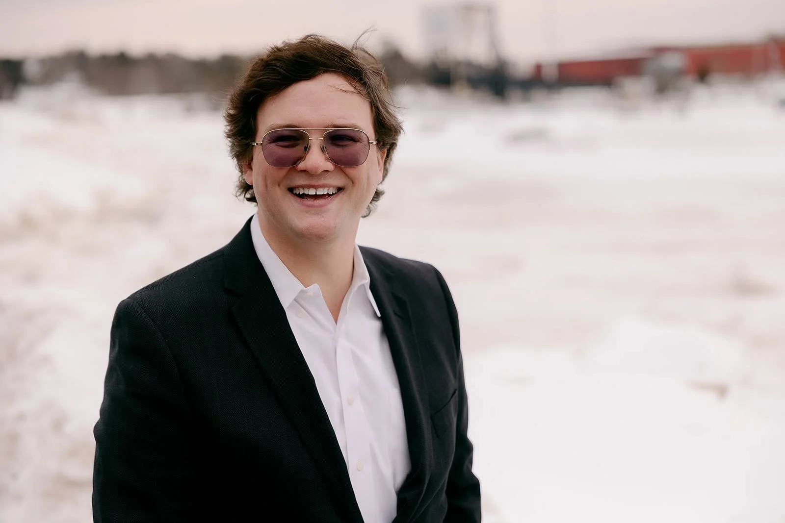 Portrait of Mike Walsh, a smiling young man with wavy brown hair and tinted aviator sunglasses, wearing a dark blazer over a white open-collar shirt, in a snowy outdoor setting.
