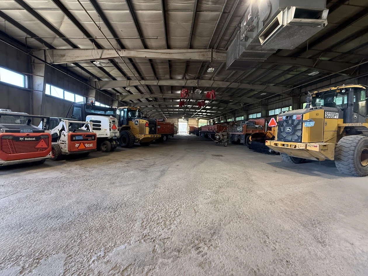 A long equipment bay at the Reading DPW filled with vehicles, including two orange Bobcat skid-steers, a white dump truck, and yellow John Deere front-end loaders parked along both sides.