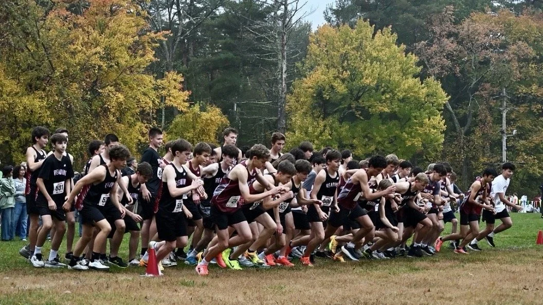 A large group of boys in black and maroon jerseys surges off the starting line on a grass field in front of autumn trees.