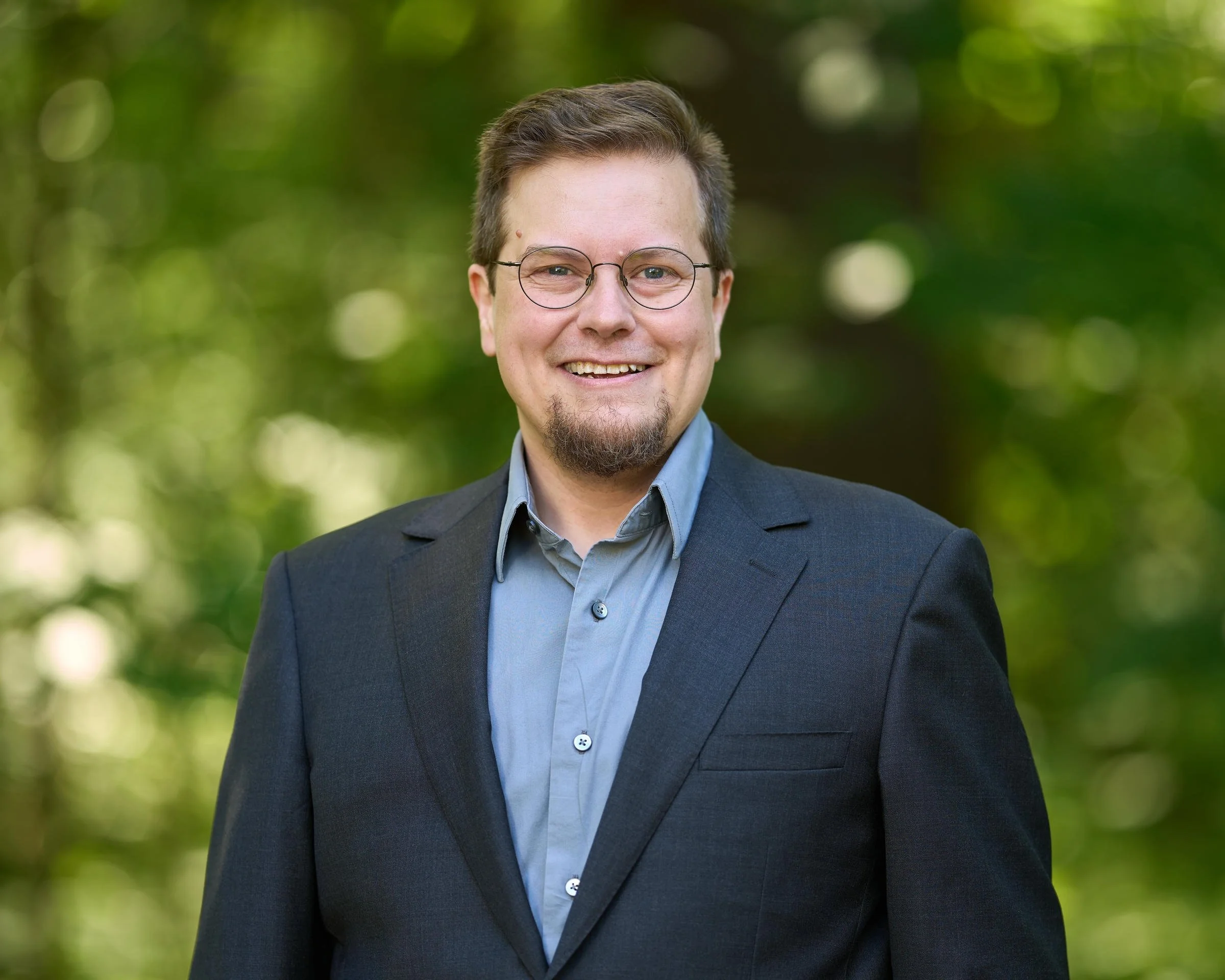Campaign photo of Beth Andres-Beck, a person with short brown hair, round glasses, and a goatee, smiling outdoors against a leafy green background, wearing a dark gray suit jacket over a blue shirt.