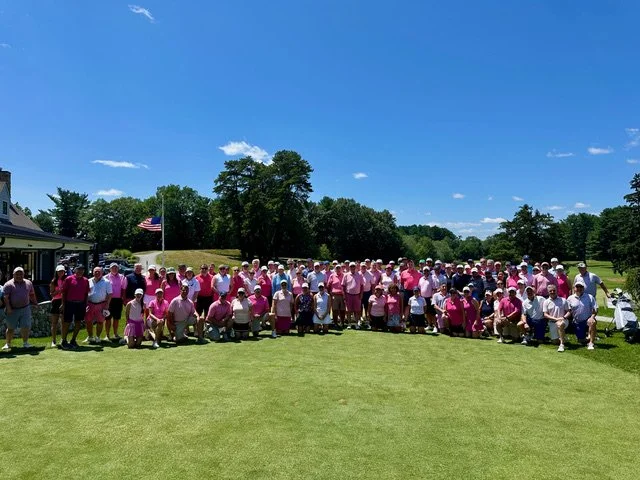 A large group of golfers and volunteers dressed in pink pose together on a putting green at Meadow Brook Golf Club under a clear blue sky, with the clubhouse, an American flag, and tree-lined fairway in the background.
