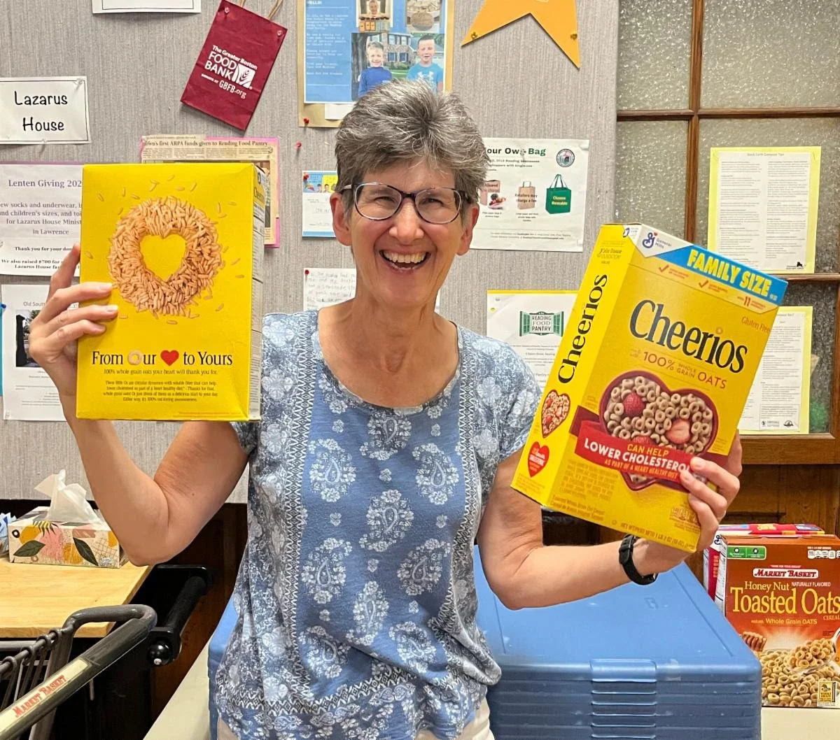 A smiling volunteer holds two boxes of Cheerios at the Reading Food Pantry.
