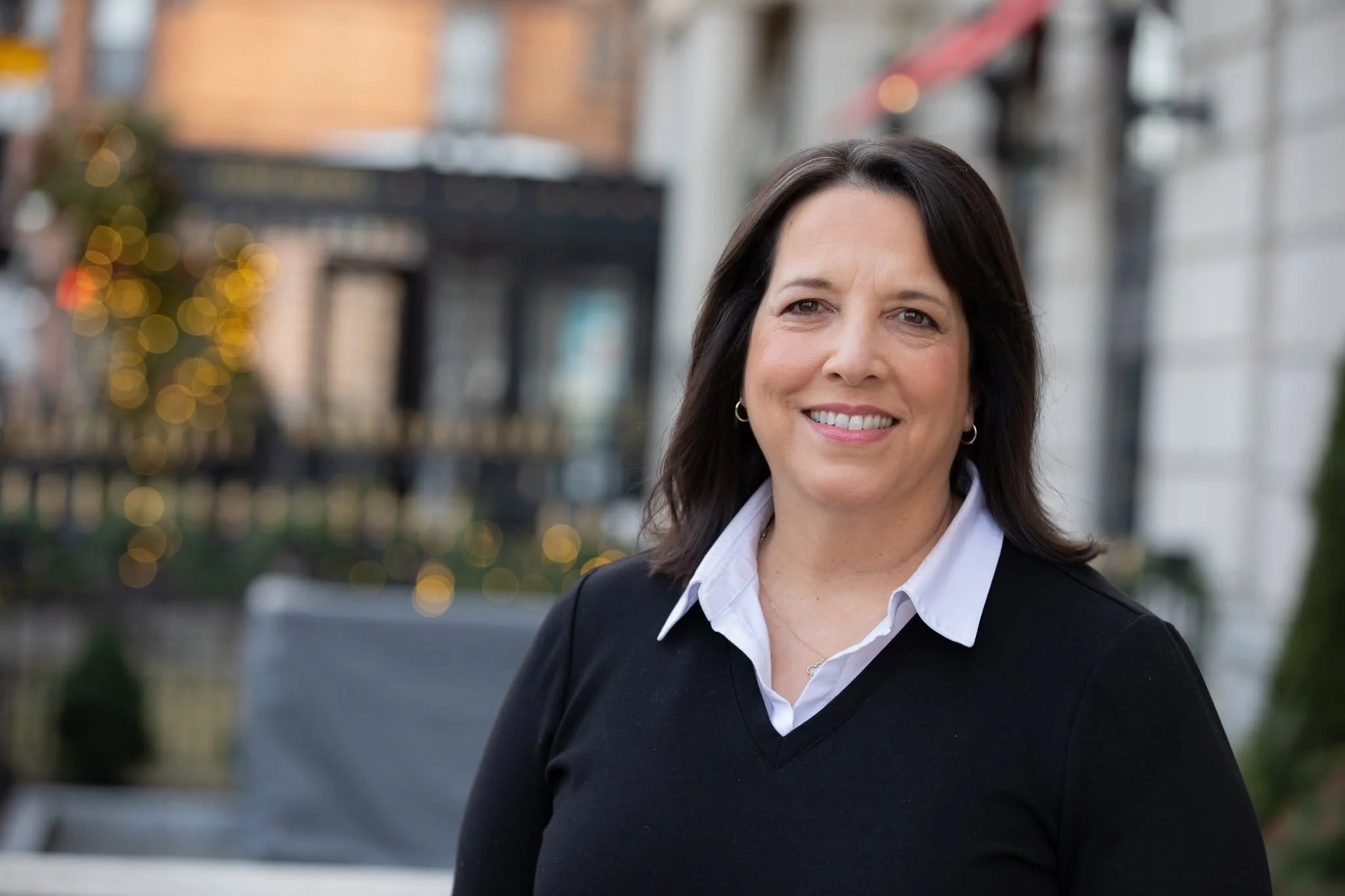 Portrait of Kim Driscoll, a middle-aged white woman with dark shoulder-length hair in a black V-neck sweater over a white collared shirt, on a city street.