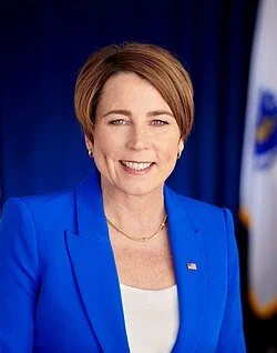 Official portrait of Governor Maura Healey, a middle-aged white woman with short reddish-brown hair, wearing a bright blue blazer over a white top, standing in front of a dark background with a flag.