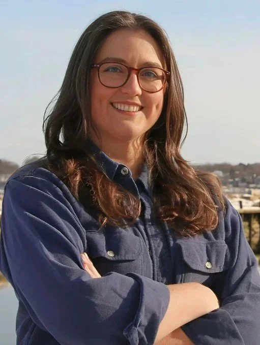 Portrait of Mariah Lancaster, a smiling woman with long brown hair and tortoiseshell glasses, arms crossed, wearing a navy button-up jacket, at a waterfront.