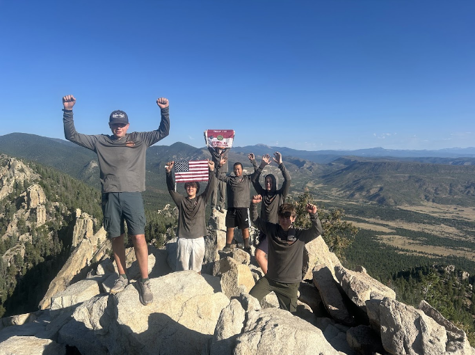 Five hikers on a rocky summit raise their arms, one pair holding a U.S. flag and another a red and white flag, with forested mountains and blue sky behind.