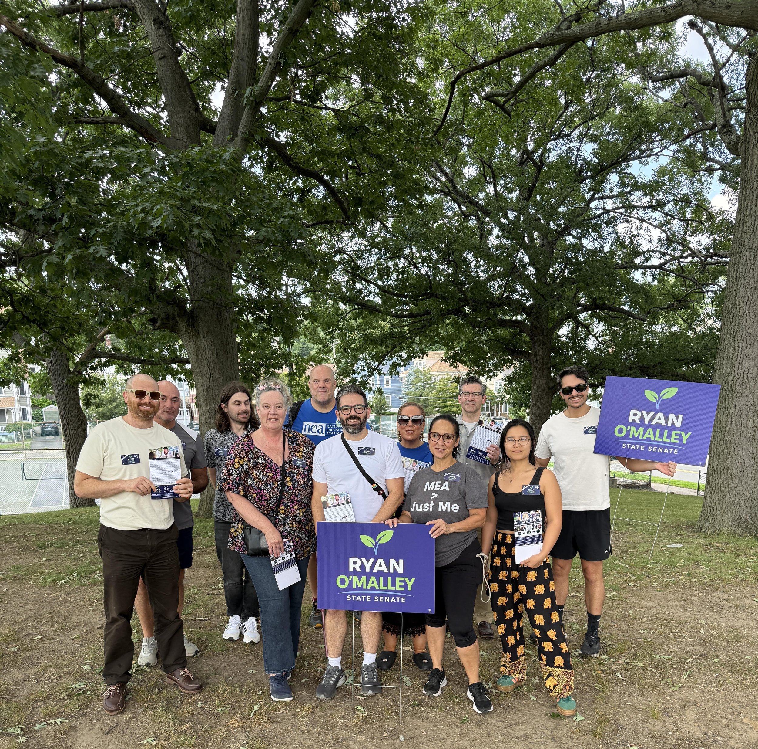A group of about ten volunteers and supporters pose outdoors under trees, several holding "Ryan O'Malley State Senate" yard signs, some wearing NEA and MTA-affiliated shirts.