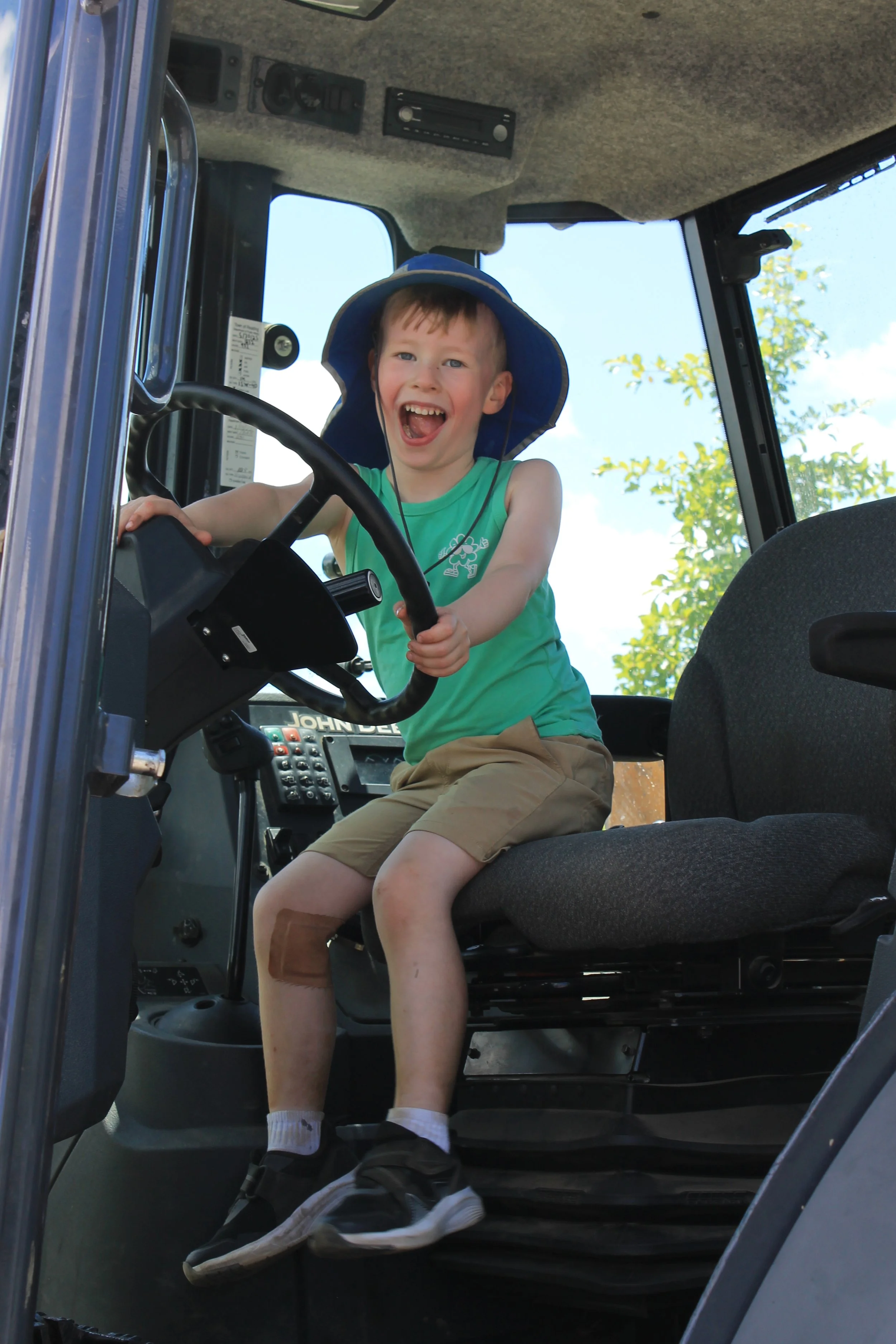 A grinning child in a green tank top and blue sun hat sits in the driver's seat of a John Deere vehicle, gripping the steering wheel with both hands.