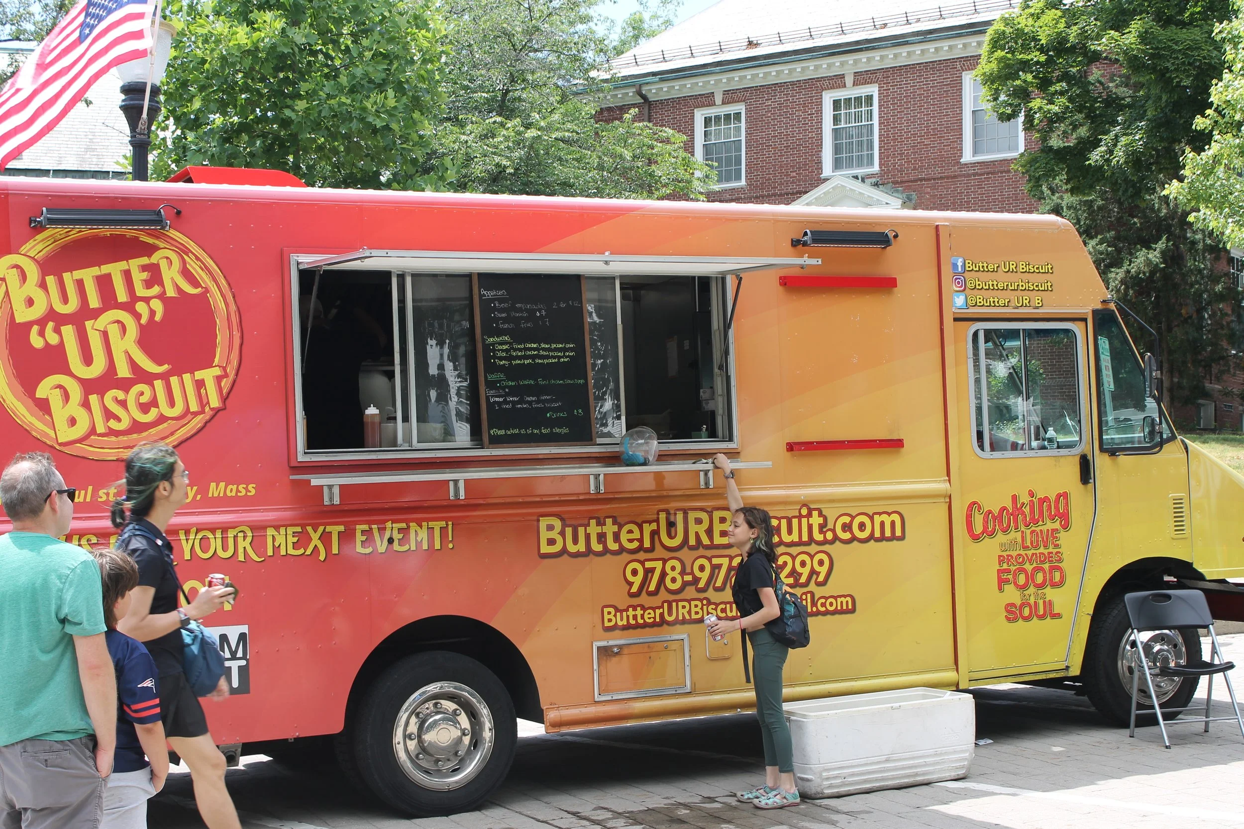 A red, orange, and yellow food truck labeled Butter "UR" Biscuit parked on a brick plaza, with a chalkboard menu in the service window and people waiting nearby.