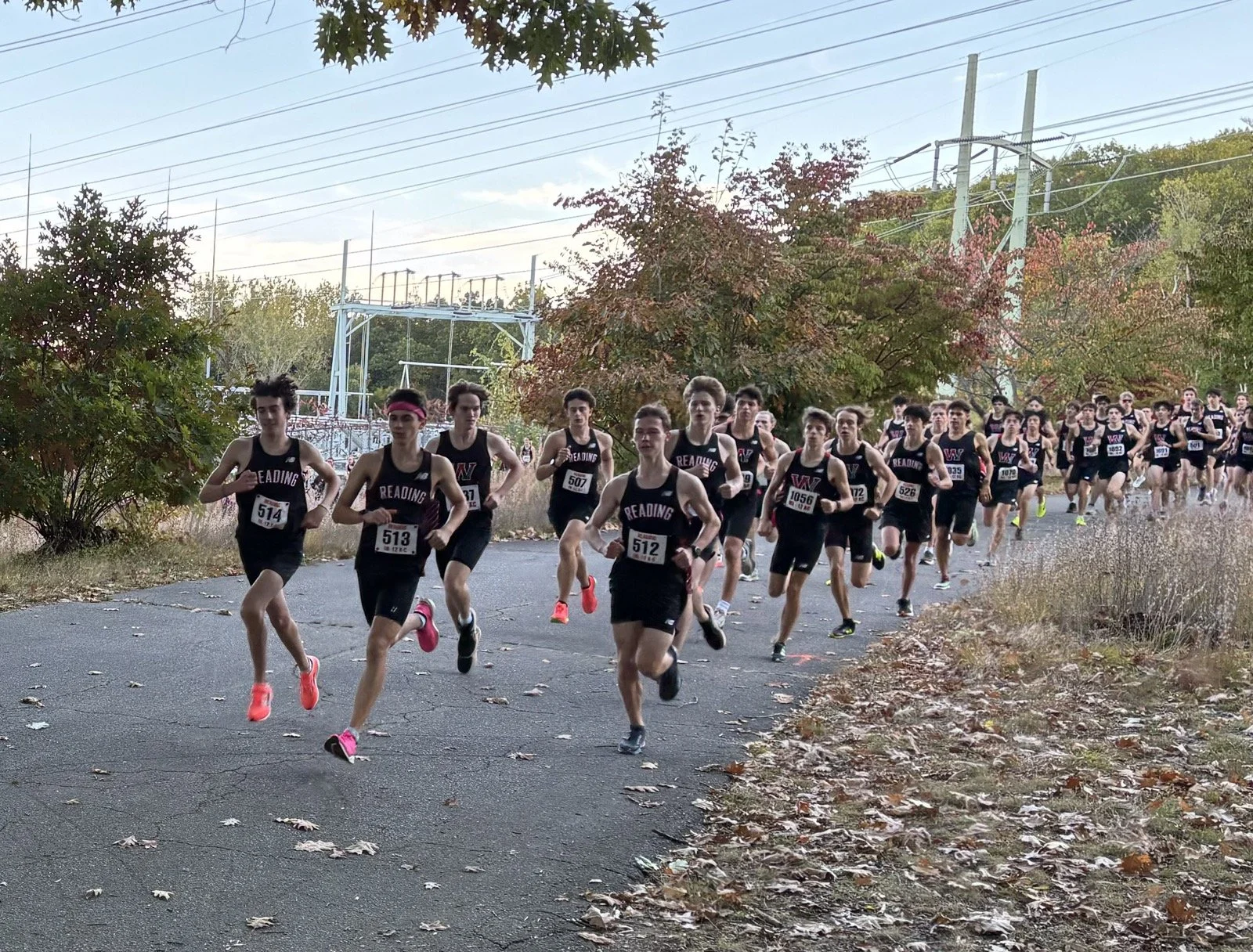 Reading boys cross-country runners lead a large pack at the start of a race on a path near power lines, with Winchester runners close behind.