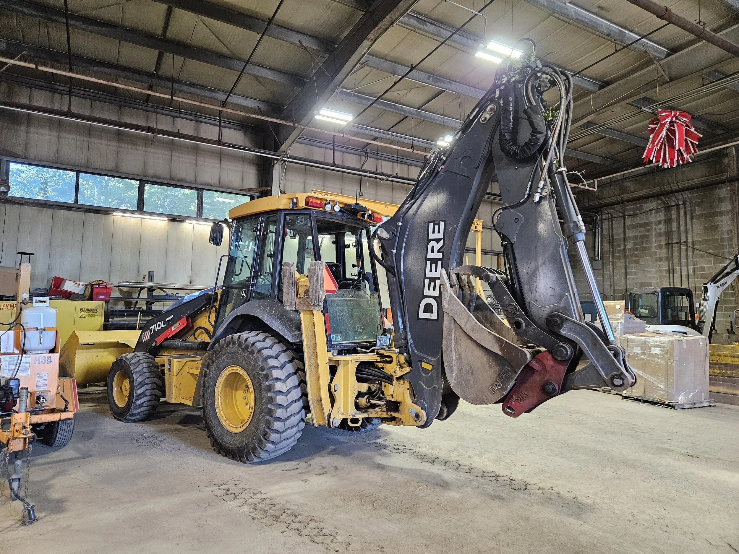 A yellow John Deere 710L backhoe with its arm and bucket raised, parked inside a garage.