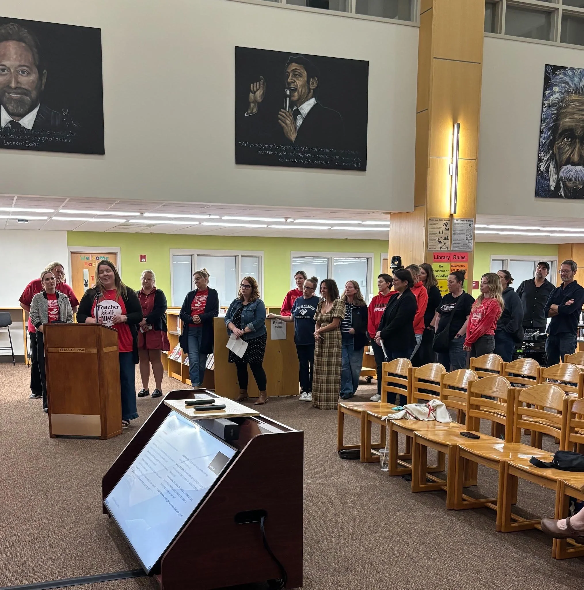 A teacher in a red shirt speaks at a wooden podium in a school library as a group of educators, many wearing red, stand behind her. Rows of wooden chairs sit empty at right.
