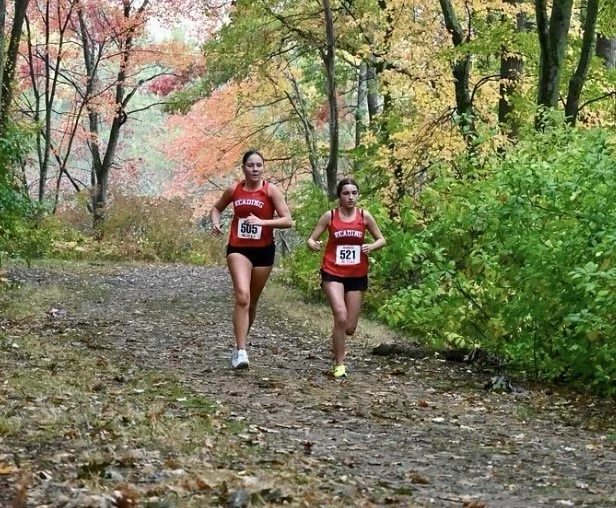Two Reading girls in red uniforms, bibs 505 and 521, run along a leaf-covered trail lined with colorful fall foliage.