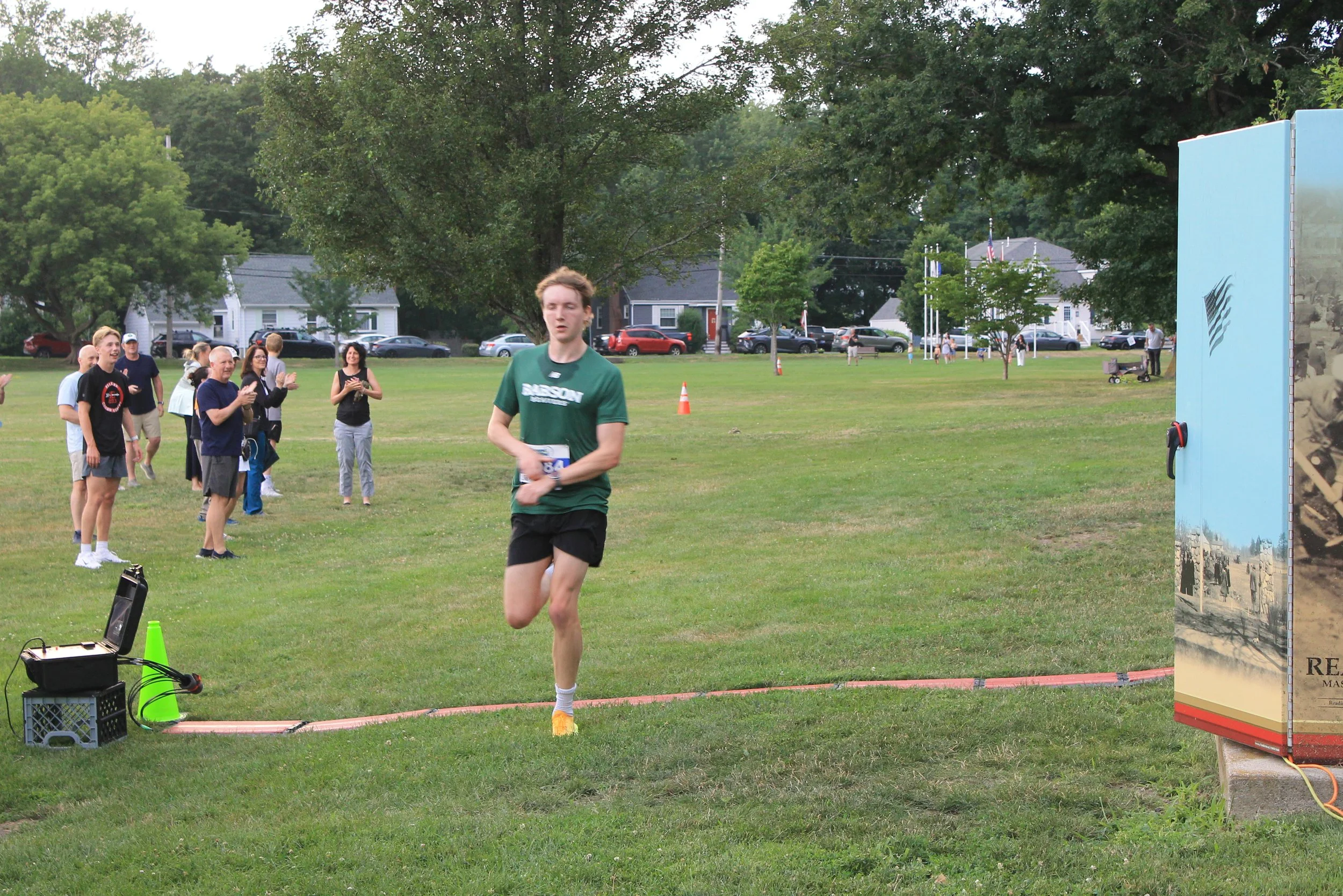 A runner in a green Babson shirt and black shorts approaches the finish line on a grassy field as a small crowd applauds from the sideline.