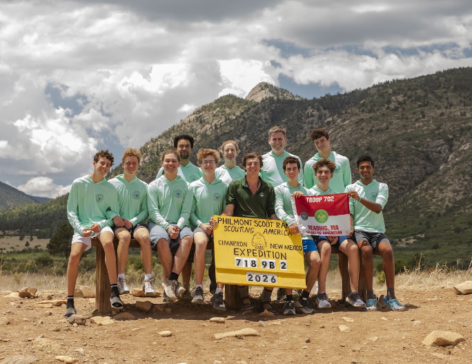 Group in light green shirts sits and stands behind a yellow Philmont Scout Ranch Cimarron, New Mexico Expedition sign reading 718 9B 2 2026 and a Troop 702 Reading, MA flag.