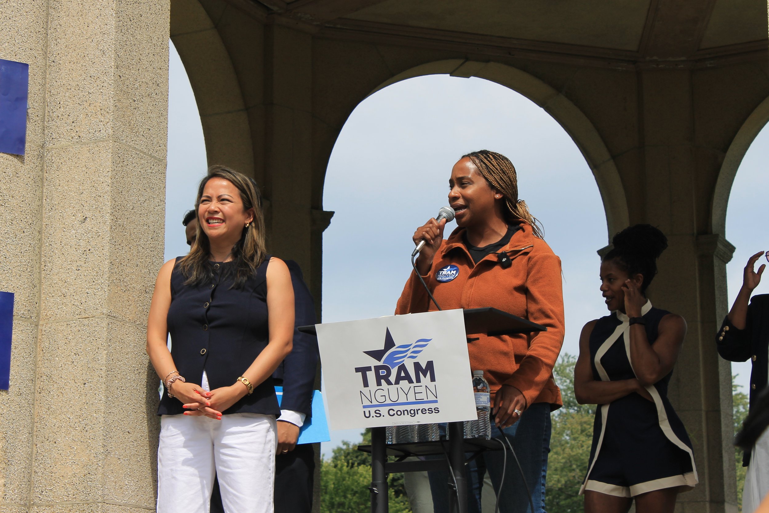 A speaker with braided hair, wearing an orange jacket, speaks into a microphone beside Tram Nguyen, with supporters clapping.