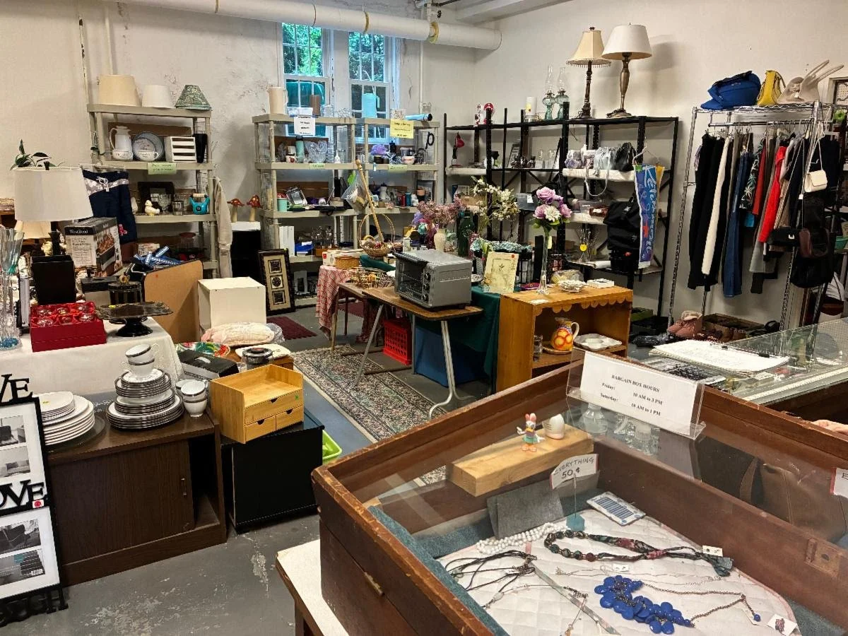 Interior of the Bargain Box thrift shop, with shelves of housewares, glassware, lamps, and clothing racks, plus a display case of jewelry. A sign lists hours as Friday 10 a.m. to 3 p.m. and Saturday 10 a.m. to 1 p.m.