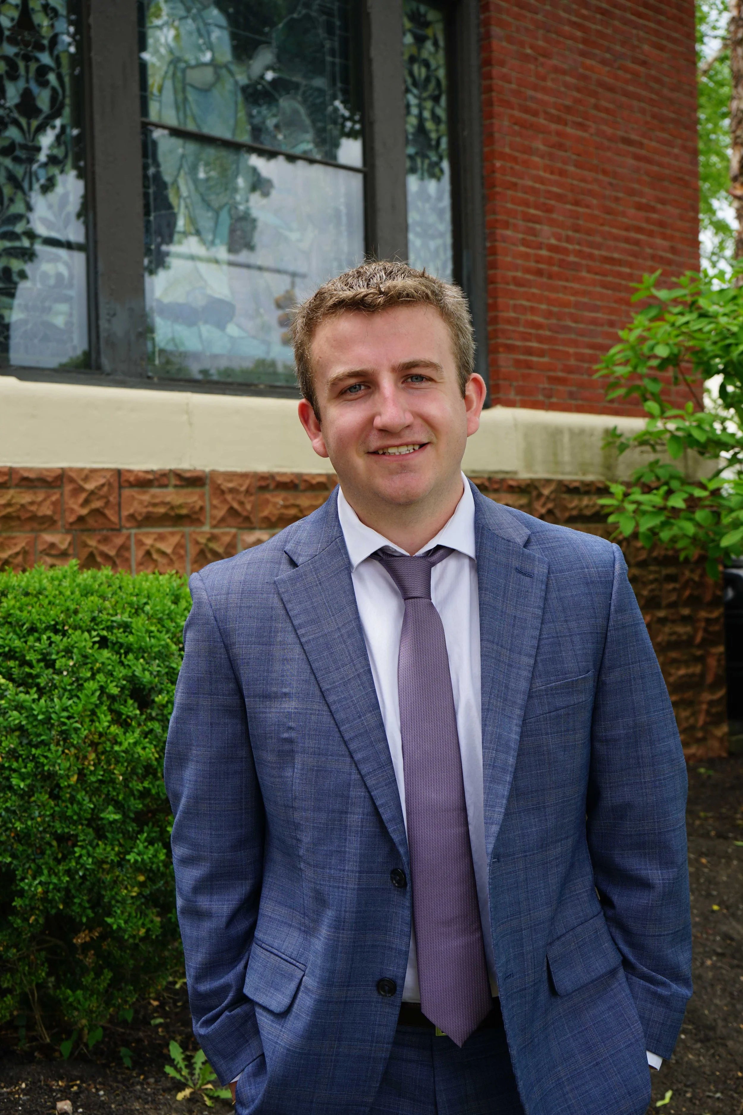 Portrait of Trevor Currier, a smiling young man in a blue plaid suit with a mauve tie, outside a brick building.