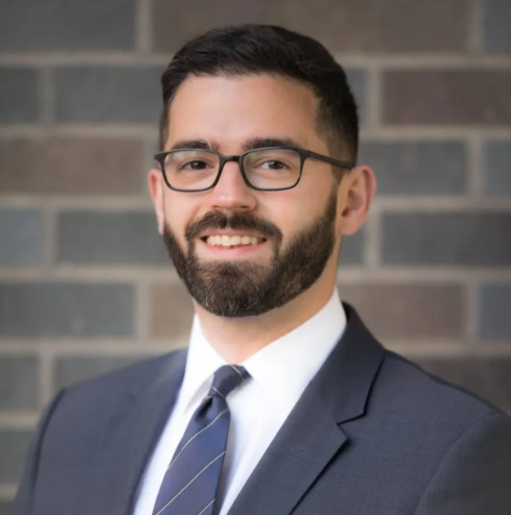 Portrait of Ryan O'Malley, a smiling young man with dark hair, a beard, and glasses, wearing a navy suit with a white shirt and striped tie, against a gray brick wall.