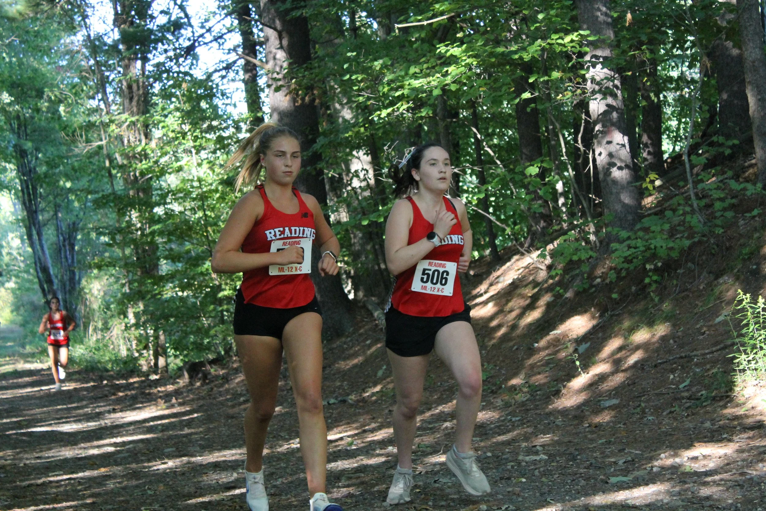 Two Reading girls cross-country runners run side by side on a wooded trail during a race.