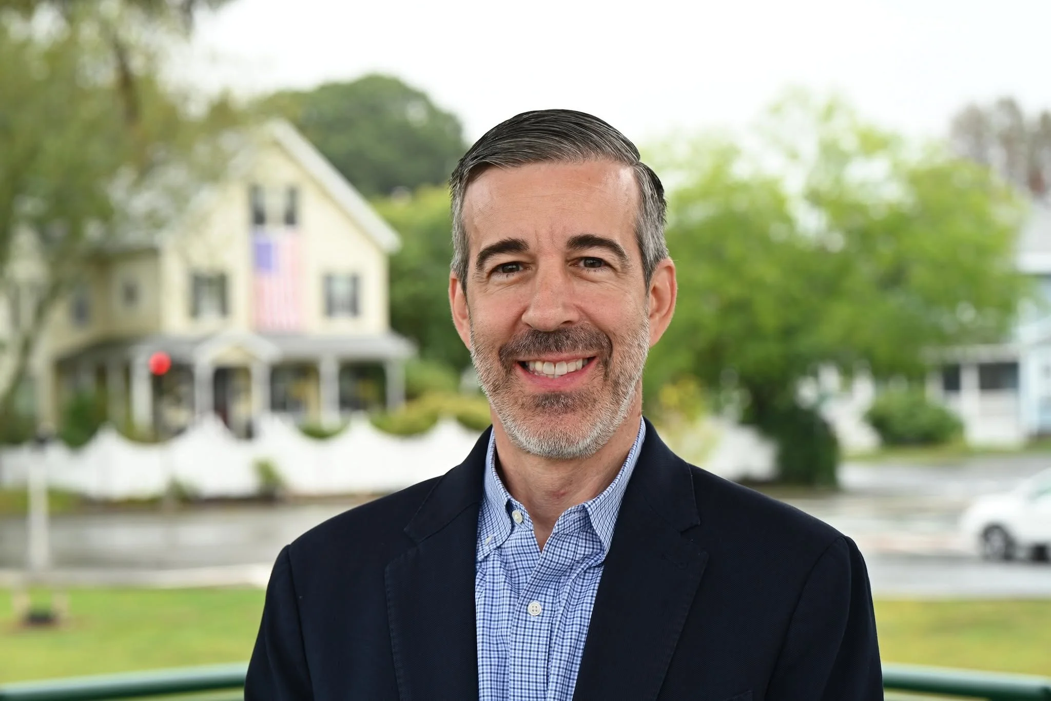 Portrait of John Beccia, a smiling middle-aged white man with graying hair and a beard, wearing a navy blazer over a blue checked shirt, with a house and American flag behind him.