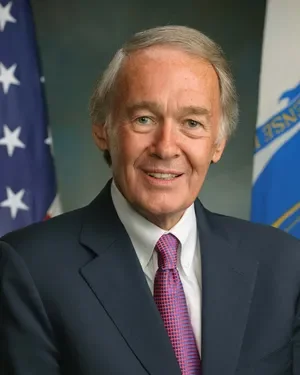 Official portrait of Senator Ed Markey, an older white man with gray hair, wearing a dark suit and purple tie, standing in front of an American flag and the Massachusetts state flag.