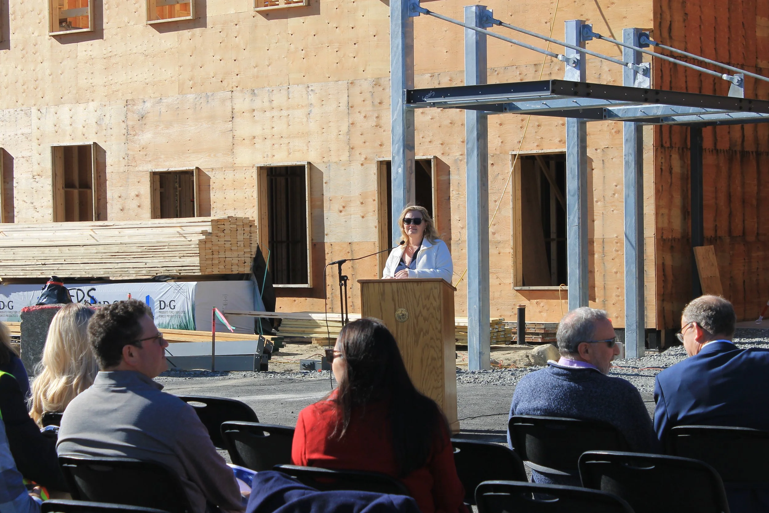 Woman with blond hair and sunglasses speaks at a wooden podium before the wood-framed building, with seated attendees and stacked lumber nearby.