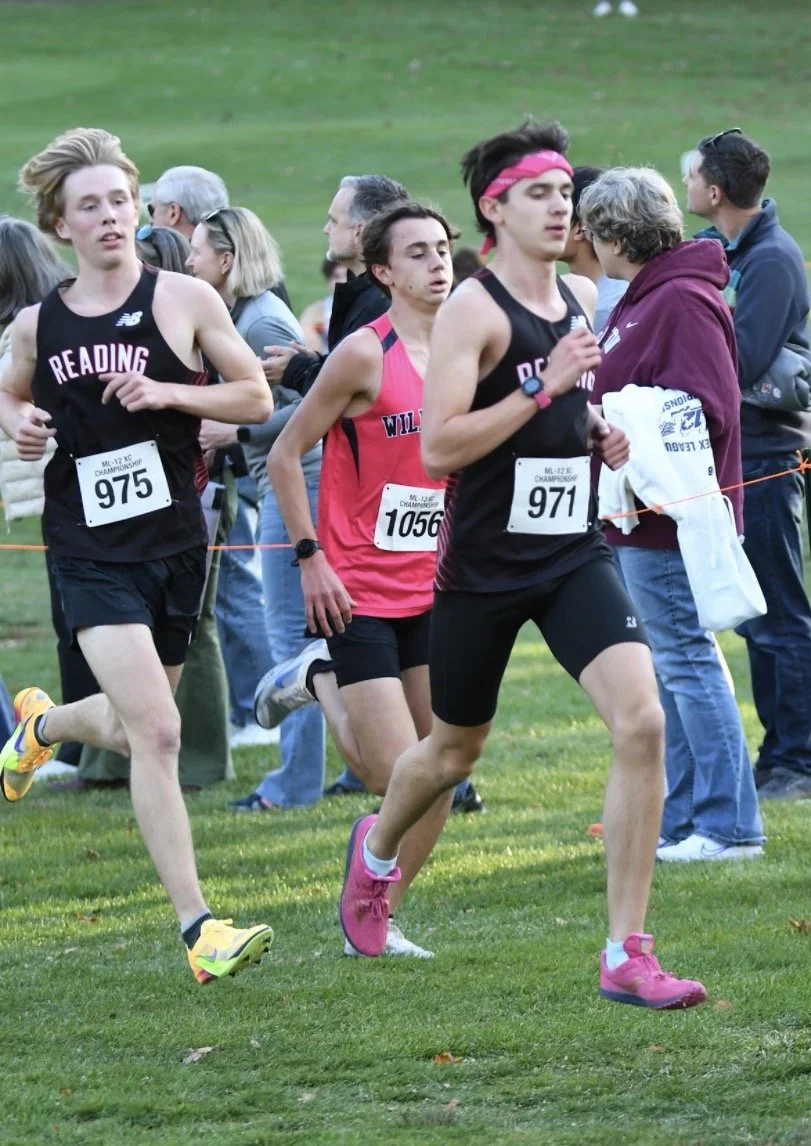 Two Reading boys runners in black uniforms, bibs 975 and 971, one wearing a pink headband, run alongside a Wilmington runner in pink as spectators watch from behind a rope.