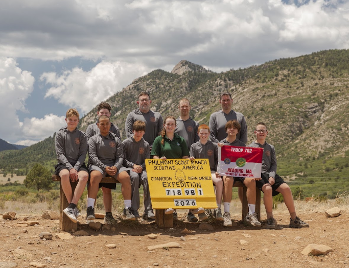 Group in gray shirts sits and stands behind a yellow Philmont Scout Ranch Cimarron, New Mexico Expedition sign reading 718 9B 1 2026 and a Troop 702 Reading, MA flag, with mountains behind.