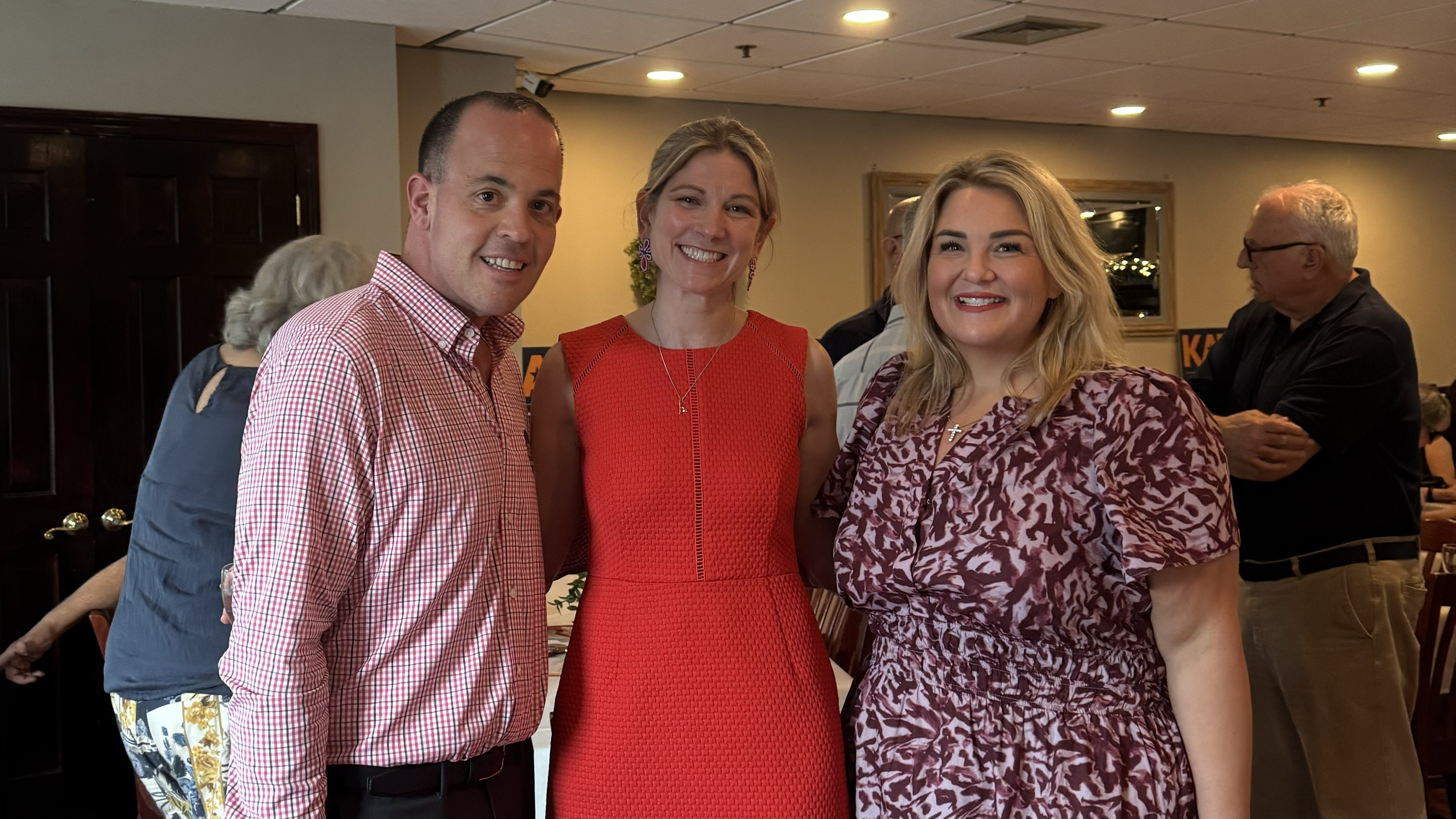 Kate Lipper-Garabedian, in a red dress, stands between a man in a checked shirt and a woman in a patterned dress at an indoor campaign event.