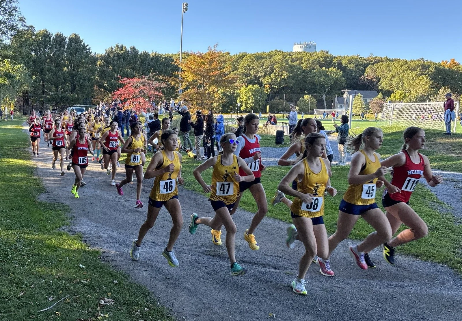 Girls at the start of a cross-country race, in red Reading and yellow uniforms, run down a gravel path past spectators with autumn trees and a field behind them.