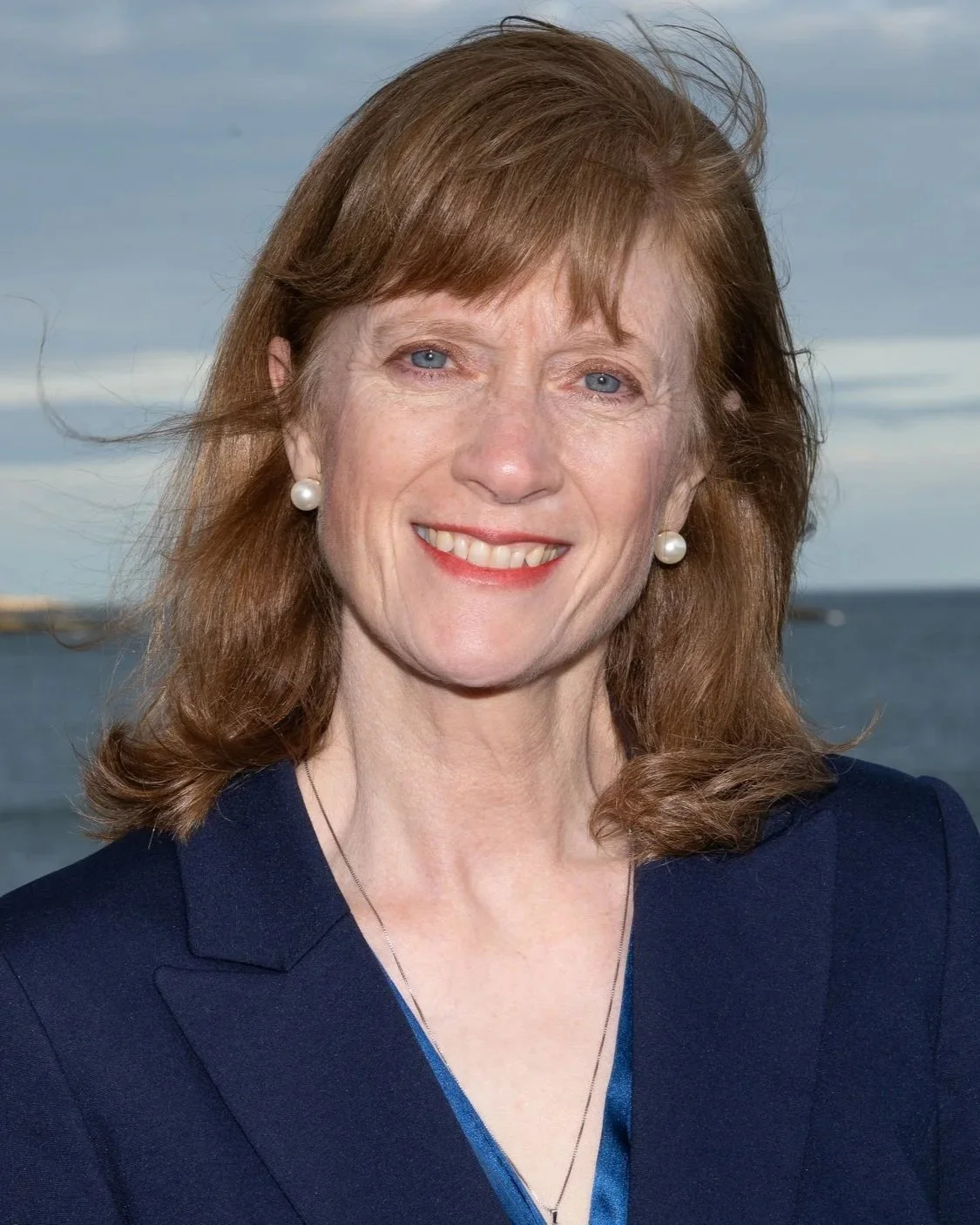 Portrait of Diann Baylis, a smiling woman with shoulder-length reddish-brown hair and pearl earrings, wearing a navy blazer over a blue top, at a rocky ocean shoreline.