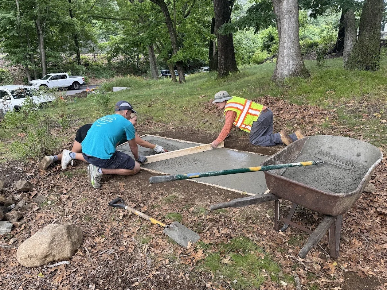 Three people kneel on either side of a freshly poured concrete pad, smoothing the surface with a board, with a wheelbarrow of concrete nearby.