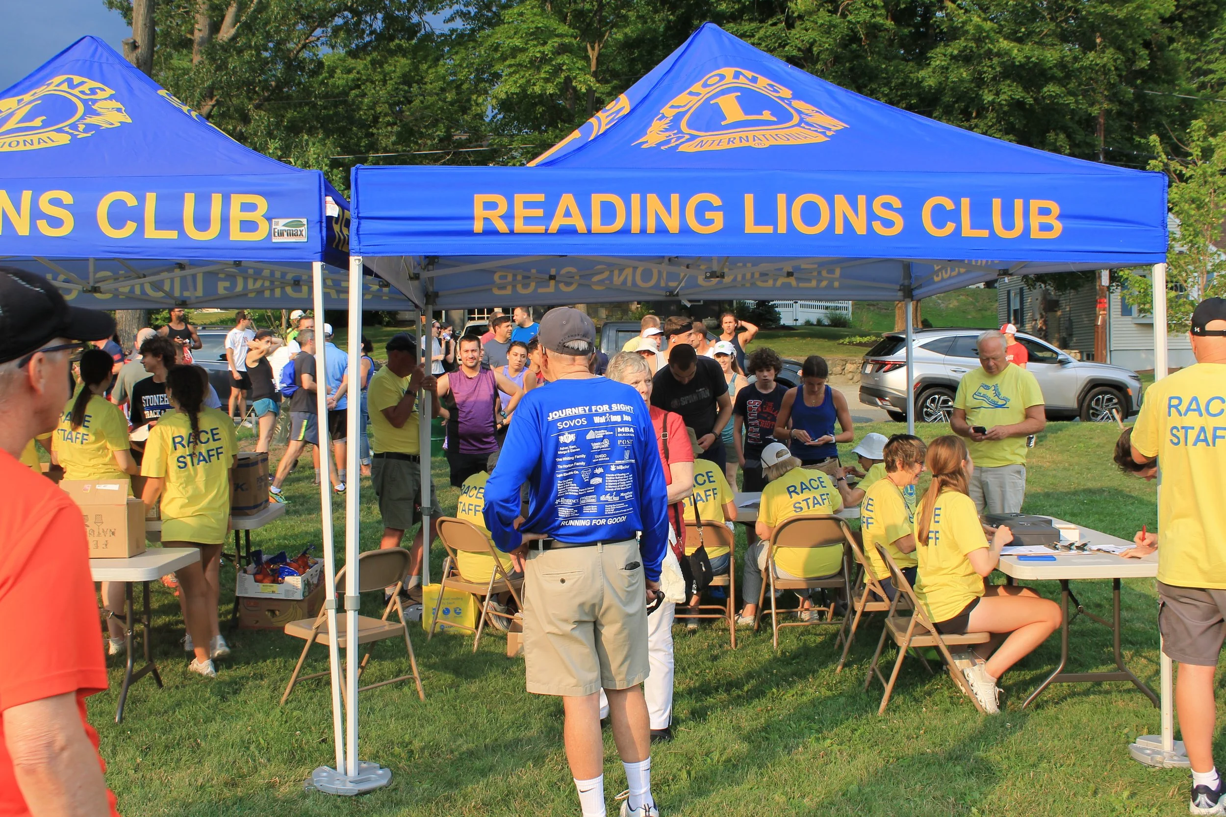 Runners gather at check-in tables staffed by volunteers in yellow "Race Staff" shirts, under two blue tents reading "Reading Lions Club."
