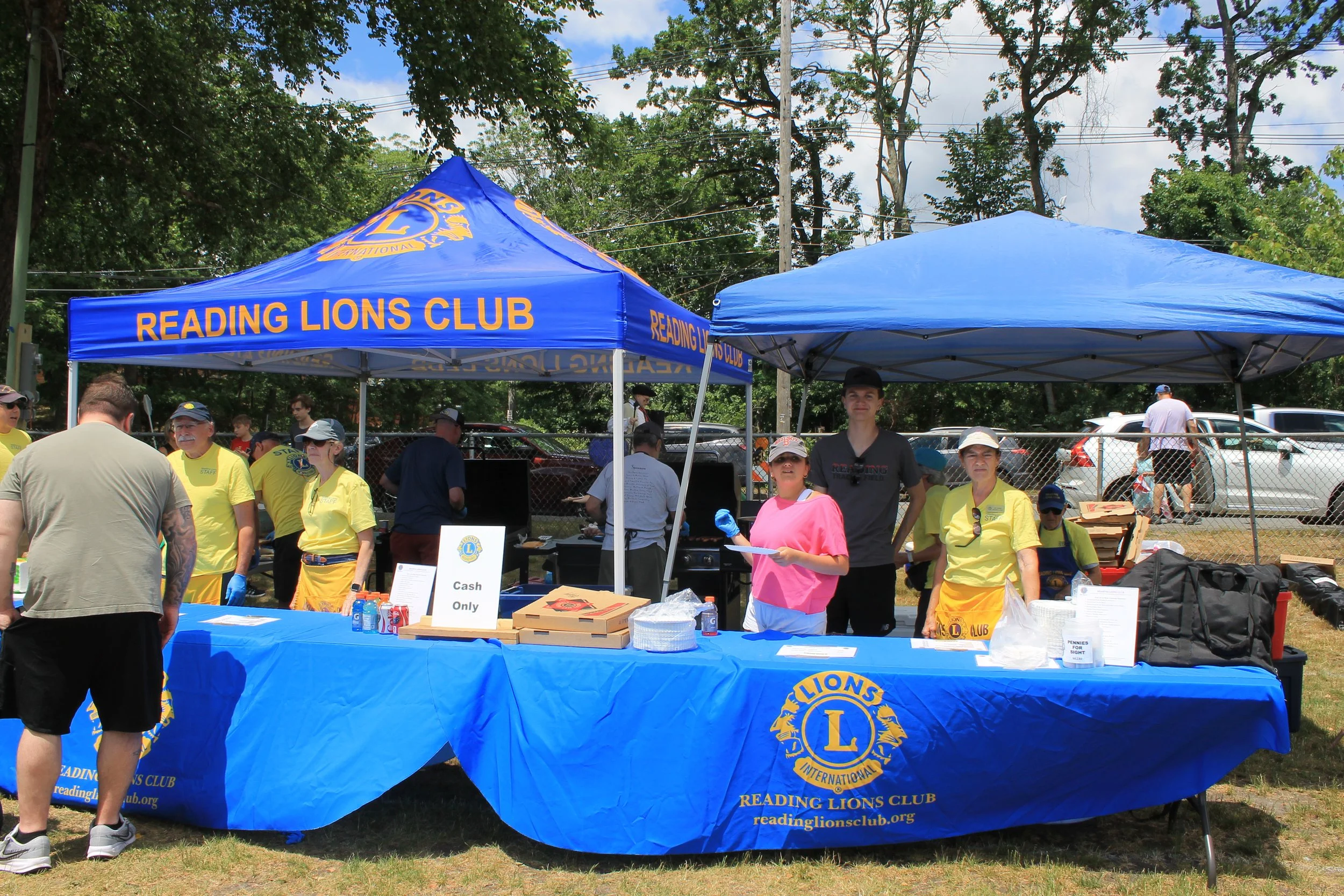 Volunteers in yellow Lions Club shirts staff a food table under a blue Reading Lions Club tent, with pizza boxes, drinks, and a grill set up behind them.