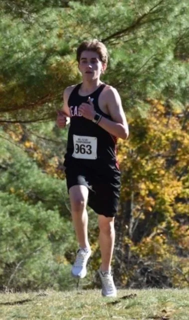 A Reading boys cross-country runner in a black uniform and bib 963 runs across a grassy hill with pine trees and fall foliage in the background.